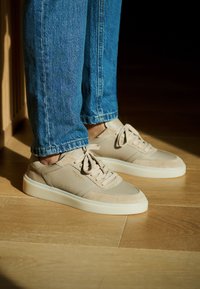 Zapatillas beige hechas de ante y cuero, con suela de goma cremosa, cordones y detalles perforados sutiles en los laterales.