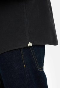 Maglietta nera con texture a coste, caratterizzata da un orlo curvo. Piccola etichetta bianca con il nome del marchio visibile in basso, abbinata a jeans in denim scuro.