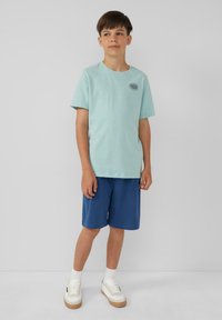 Lichtblauwe katoenen t-shirt met korte mouwen, voorzien van een ronde halslijn en een klein zwart logo. Gekleed met blauwe shorts en witte sneakers.