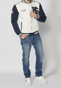 Veste varsity blanche et marine avec lettres et insignes brodés, associée à un jean bleu et des baskets blanches. Dotée de poignets et d'un ourlet côtelés.