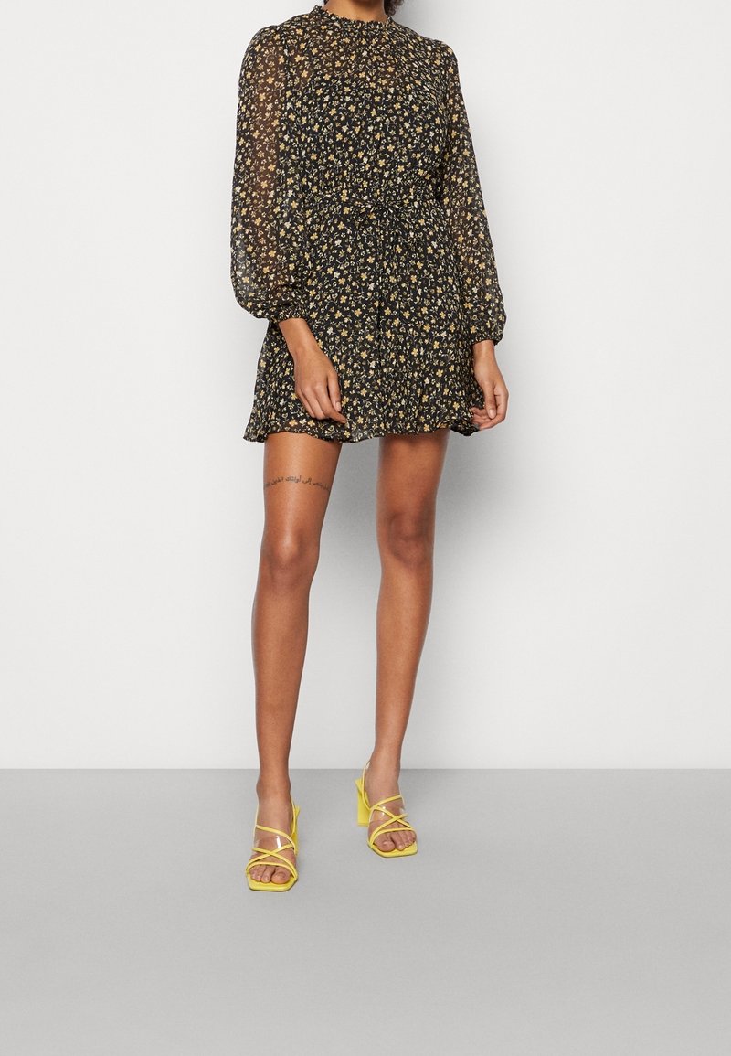 Femme portant une robe courte noire à fleurs avec des manches longues transparentes et des sandales à talons jaunes, debout devant un fond clair uni.