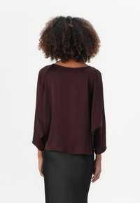 Top bordeaux fluide avec de larges manches et un col bateau, porté sur une jupe noire ajustée, présentant une texture douce et une finition lisse.