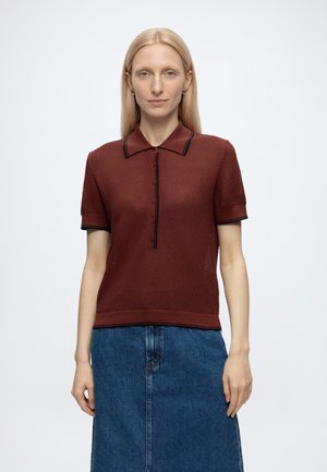 Femme aux cheveux blonds raides portant un polo bordeaux à manches courtes texturé et une jupe en jean bleue, debout devant un fond uni clair.