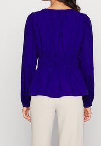 Top peplum viola con maniche lunghe, vita elasticizzata e dettagli arricciati sulle spalle. Il tessuto morbido e la forma aderente esaltano il design.