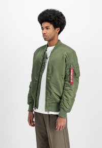 Olive green bomber jacket with a satin finish, ribbed cuffs, and waistband. Features a red accent tag on the sleeve and zippered pockets.