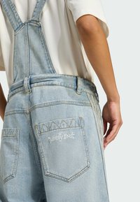 Svetlo modré denné overaly majú nastaviteľné remene, zadnú vrecko s bielym vyšívaným logom a kontrastné prešívanie na švoch.