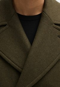 Olivgrön ullrock med texturerad yta, har en bred krage och en svart tröja under, delvis synlig i bilden.