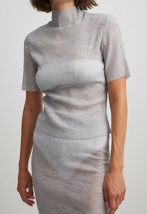 Femme portant un haut texturé gris clair à manches courtes avec col montant et une jupe assortie sur un fond clair uni.