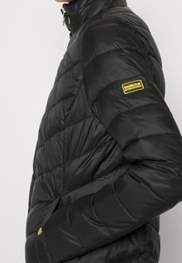 Schwarze Daunenjacke mit einem hochschließenden Reißverschlusskragen, gestepptem Design und einem gelben Barbour International-Logo auf der linken Brust.