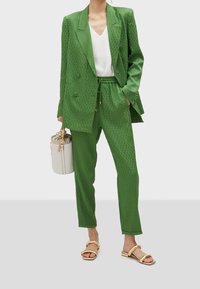 Woman wearing a green patterned oversized blazer and matching drawstring pants, white top, gold sandals, holding a white bucket bag.