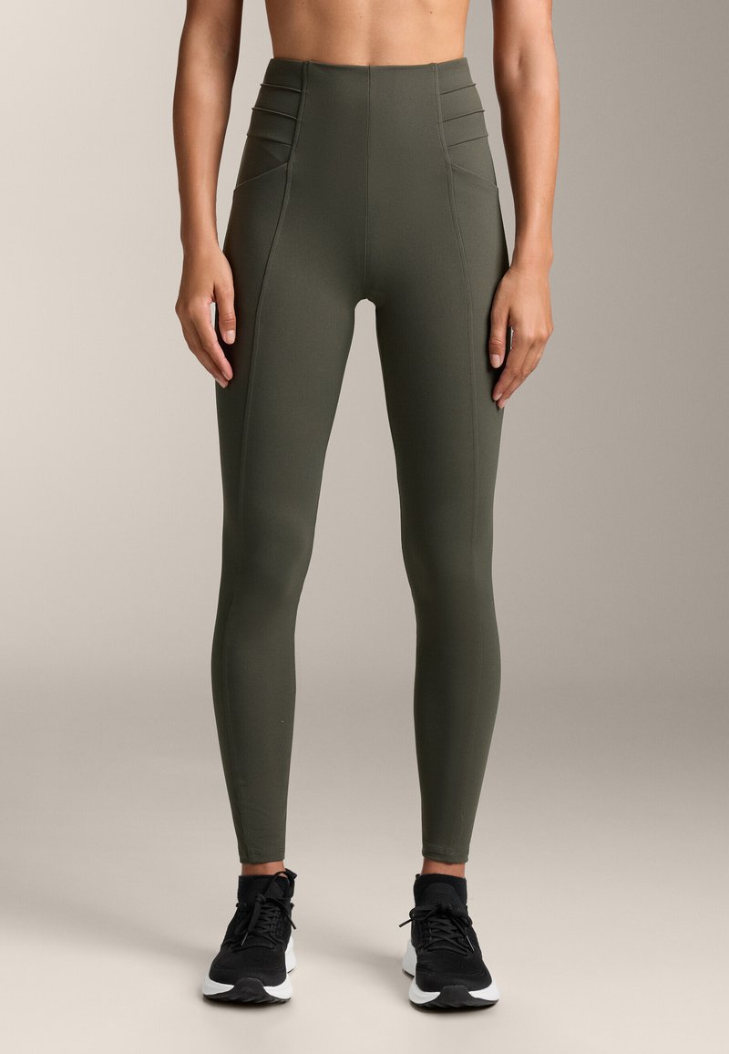 Leggings verde oliva a vita alta con gambe affusolate. Presentano cuciture testurizzate e un tessuto liscio e elasticizzato. Abbinati a scarpe da ginnastica nere.
