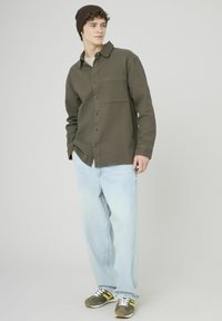 Chemise olive verte à manches longues avec un motif texturé, poche poitrine unique, associée à un jean bleu clair et des baskets brunes avec des accents jaunes.