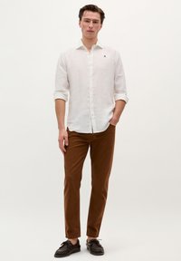 Hvid linned skjorte med knapper og optrukne ærmer, parret med brune slim-fit bukser og mørke sko. Enkel frontdesign med et lille logo.