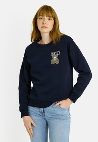 Sudadera azul marino con puños y dobladillo acanalados. Presenta el texto "PRETTY" y un diseño bordado de un oso de peluche en el pecho.