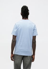 Světle modré bavlněné tričko s kulatým výstřihem a krátkými rukávy, pohled zezadu, ve spojení se šedými džínami.