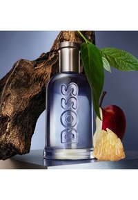 Blauwgetinte glazen parfumfles met zilveren dop met het label "BOSS" naast hout, groene bladeren, een rode appel en een stuk citrusvrucht.