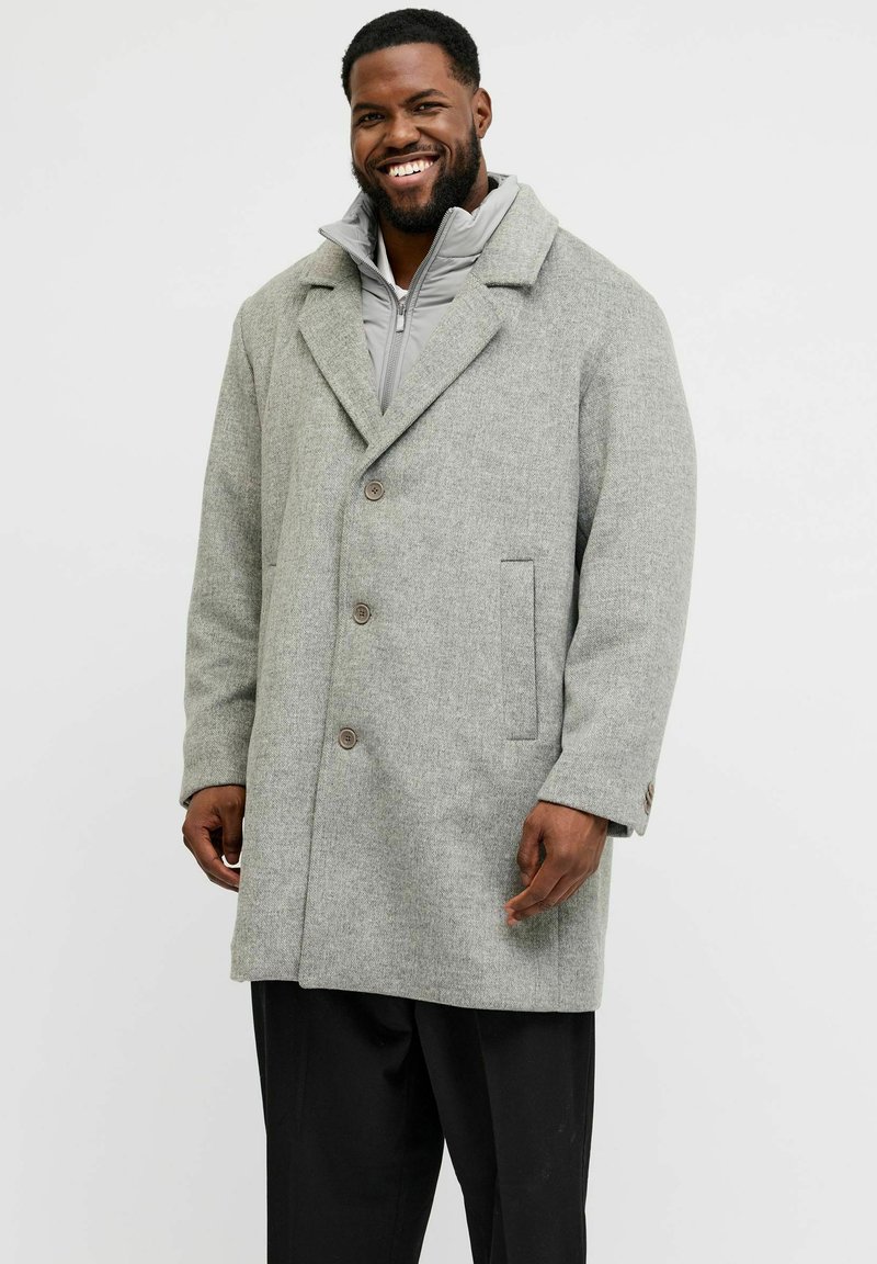 Hombre sonriente que lleva un abrigo de invierno gris claro abotonado sobre una chaqueta gris con cremallera y pantalones negros, de pie frente a un fondo liso claro.