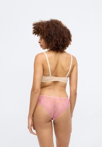 Beige BH mit verstellbaren Trägern, kombiniert mit einem rosa Spitzen-Tanga mit floralem Muster und transparentem Rücken. Glatte Stofftextur über die gesamte Fläche.
