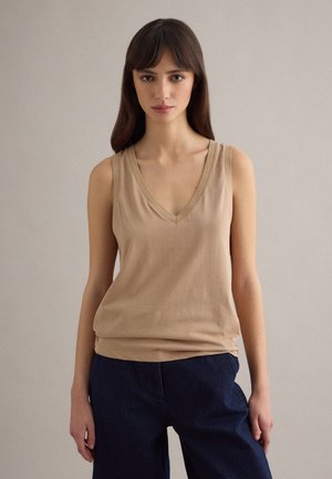 Jonge vrouw met lang bruin haar, een beige mouwloze V-hals top en donkerblauwe broek dragend, staand tegen een neutrale achtergrond.