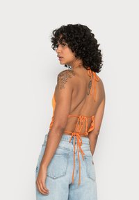 Top halter em satin laranja com design de amarração nas costas; apresenta um decote baixo nas costas e alças finas, combinado com jeans de denim claro de cintura alta.