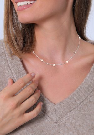 Silver necklace with small, round, rose-gold accents, featuring a delicate chain design. Worn with a beige V-neck sweater.