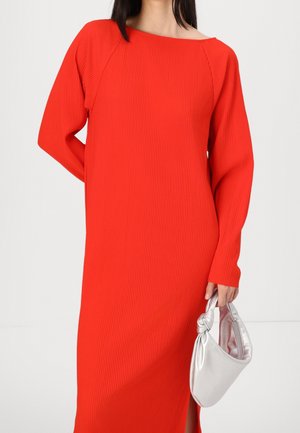 Frau in einem langärmligen, gerippten, leuchtend roten Kleid, die eine kleine, glänzende silberne Handtasche vor einem schlichten weißen Hintergrund hält.