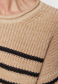 Beżowy sweter dzianinowy o teksturowej powierzchni, z poziomymi czarnymi paskami i okrągłym, prążkowanym dekoltem.