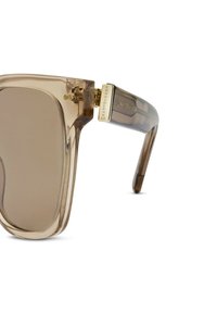 Průhledné oversized sluneční brýle vyrobené z lehkého acetátu s hnědým nádechem a zlatými akcenty na spáncích. Hladký povrch a úhlový design.