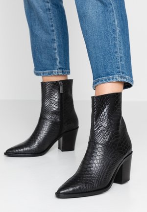 Bottines noires effet peau de serpent avec talons blocs portées avec un jean bleu retroussé sur un fond clair.