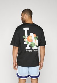 Sort T-shirt med store hvide "LA" bogstaver og et design af orange frugter og blade på ryggen. Blødt stof med en afslappet pasform.