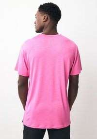 Uomo di spalle che indossa una maglietta a maniche corte rosa acceso e pantaloni neri su uno sfondo bianco semplice.