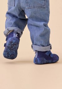 Bottes bleues pour tout-petits avec des bandes velcro, un corps texturé et une semelle à motifs. Portées avec un pantalon en denim retroussé, montrant un design décontracté.