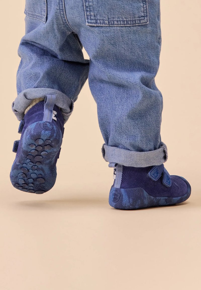 Bottes bleues pour tout-petits avec des bandes velcro, un corps texturé et une semelle à motifs. Portées avec un pantalon en denim retroussé, montrant un design décontracté.
