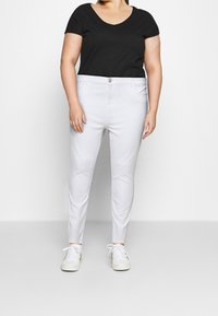 Pantalones slim-fit blancos con cierre de botón y bolsillos frontales, combinados con una blusa negra de manga corta con escote en forma de barco y zapatillas blancas.