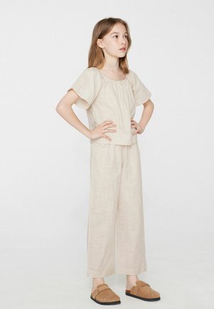 Jeune fille debout, mains sur les hanches, portant un haut beige à manches courtes, un pantalon large assorti et des chaussures marron sans lacets sur un fond uni.