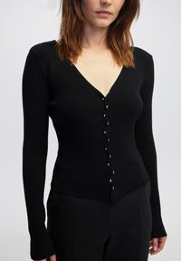 Schwarze gerippte Strickjacke mit V-Ausschnitt, langen Ärmeln und goldfarbenen Knöpfen auf der Vorderseite. Figurbetonte Form, weicher Stoff.