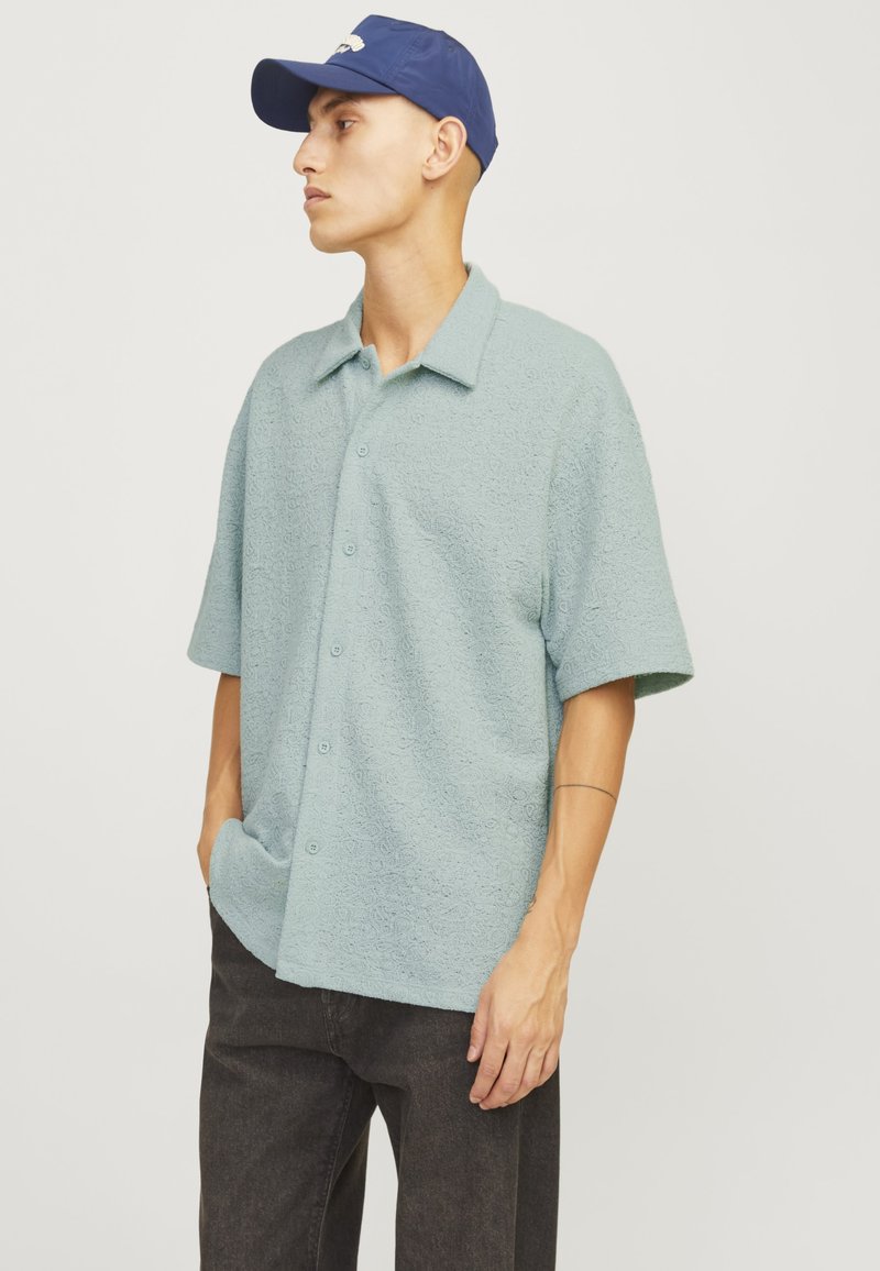 Camicia verde chiaro a maniche corte in tessuto texturizzato con motivi sottili; presenta un colletto e una chiusura con bottoni. Indossata con pantaloni scuri e un berretto blu.