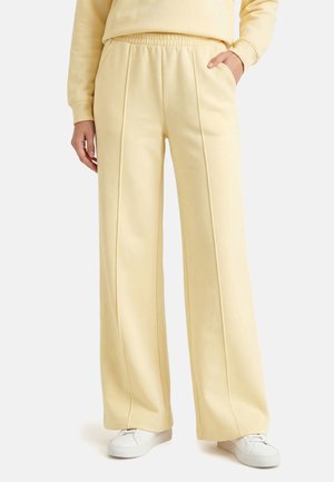 Vrouw draagt lichtgele wijde broek met voornaad, elastische taille, zijzakken, gecombineerd met witte sneakers en bijpassende top.