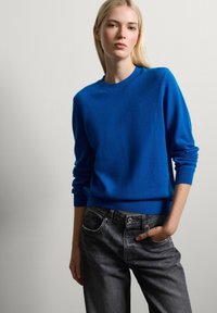 Suéter de punto azul brillante con cuello redondo y mangas largas, combinado con jeans de mezclilla gris oscuro. El suéter tiene una textura suave y puños acanalados.