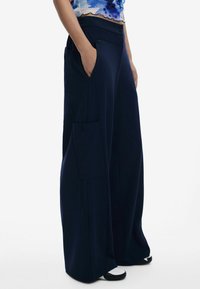 Pantaloni a gamba larga navy con tasche frontali e laterali, texture liscia, chiusura con bottone e vestibilità rilassata, abbinati a calzature bianche e nere.