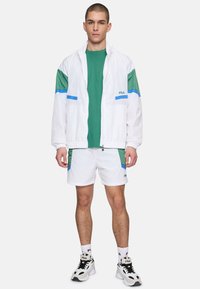 Giacca bianca con accenti verdi e blu, t-shirt verde, pantaloni corti bianchi con rifiniture verdi, sneakers nere e bianche, marchio Fila sugli indumenti.