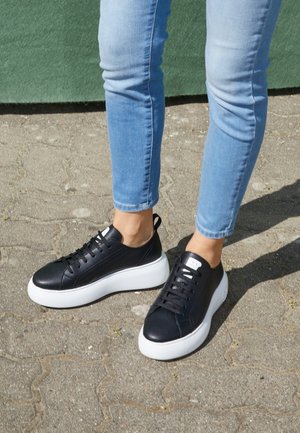 Person, die schwarze Sneakers mit dicken weißen Sohlen und hellblauen Jeans trägt, steht auf gepflastertem Boden.