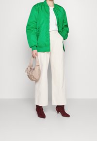Giacca bomber verde, t-shirt bianca, pantaloni larghi color crema, stivali col tacco bordeaux e una borsa in tessuto beige con manici annodati.
