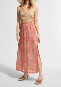 Jupe maxi avec des motifs en zigzag rouge, orange et beige. Tissu transparent, fente haute d'un côté, associée à un débardeur beige et des sandales.