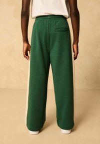 Zelené bavlnené nohavice so širokými nohavicami, s kontrastným krémovým bočným prúžkom a elastickým pásom. Obsahujú detail zadnej vrecka.