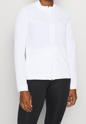 Person trägt eine weiße, langärmelige, mit Reißverschluss versehene Jacke und schwarze Hosen, steht vor einem einfarbigen hellen Hintergrund.