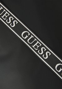 Tissu noir avec une bande diagonale de logos blancs et noirs portant le texte "GUESS", mettant en valeur une texture tissée et des couleurs contrastantes.