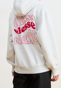 Persona che indossa una felpa leggera grigia con il logo rosso ripetuto "ellesse" sulla schiena, abbinata a pantaloni neri, mostrata da dietro.
