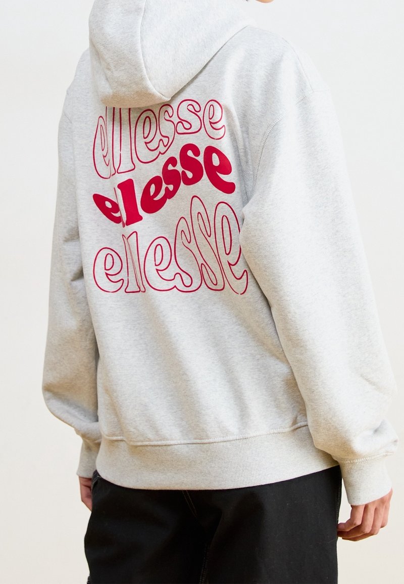 Persona che indossa una felpa leggera grigia con il logo rosso ripetuto "ellesse" sulla schiena, abbinata a pantaloni neri, mostrata da dietro.