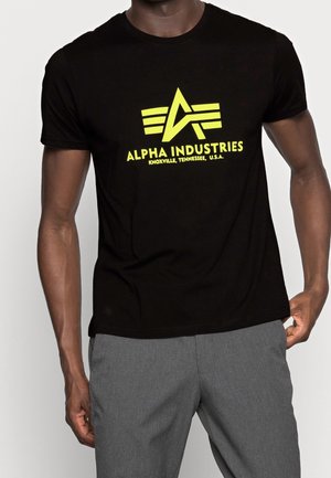 T-shirt noir avec un logo Alpha Industries jaune vif et un texte, porté par une personne en pantalon gris, debout avec une main dans la poche.